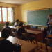 A bright, sunlit classroom with wooden desks arranged in a semicircle. Students in uniforms are attentively listening to a teacher standing near a chalkboard with Latin phrases written on it.